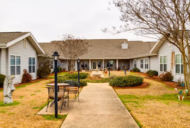 Outdoor courtyard area of a senior living facility with a concrete walkway leading to a covered patio. The courtyard features trimmed bushes, a small round table with chairs, a lamppost, and a stone angel statue on the left. The building surrounding the courtyard has light-colored siding and multiple windows.