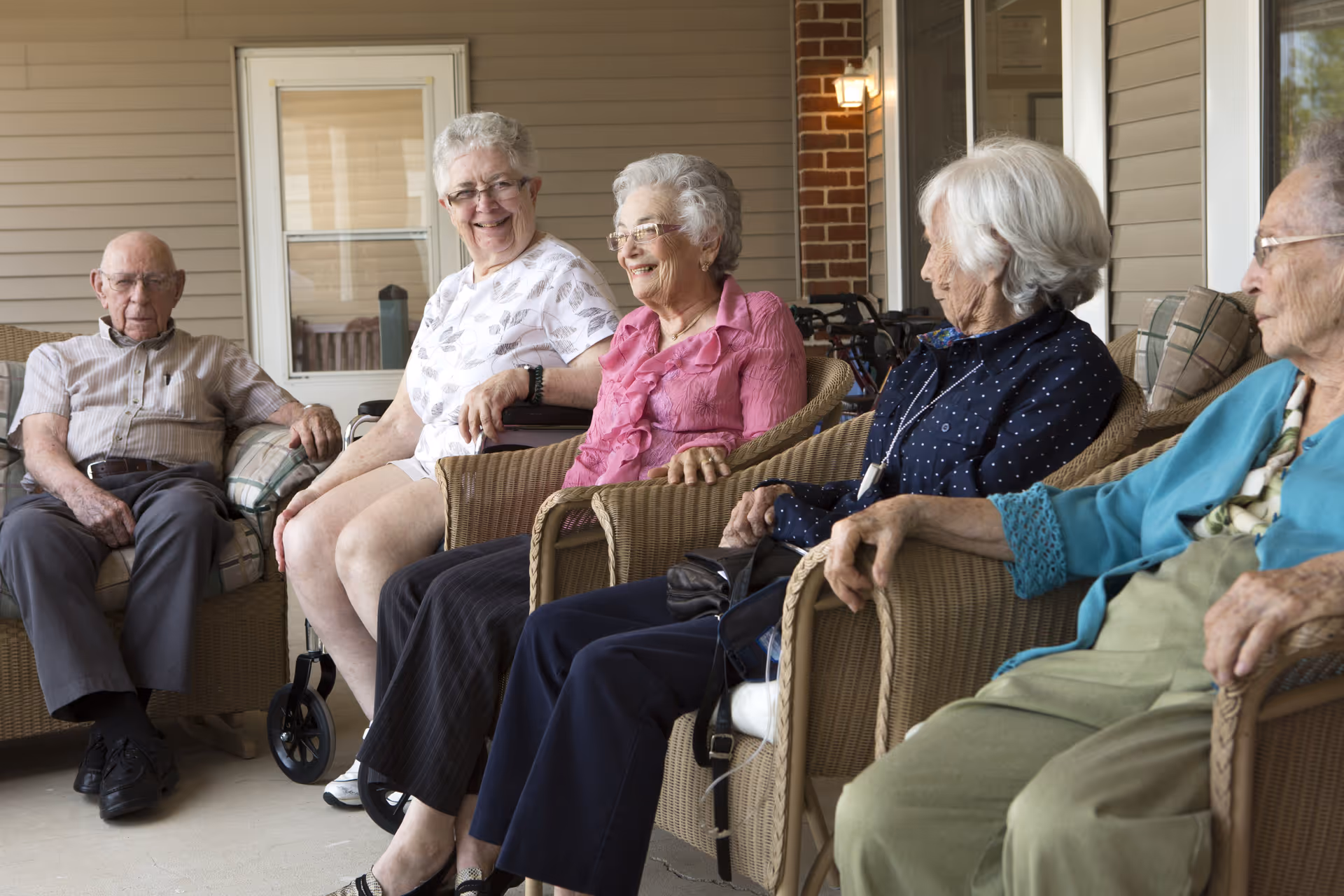 Five elderly people sitting on wicker chairs on a covered porch, engaged in conversation and smiling. The porch has beige siding, a door with a window, and a brick column. A walker is visible in the background.