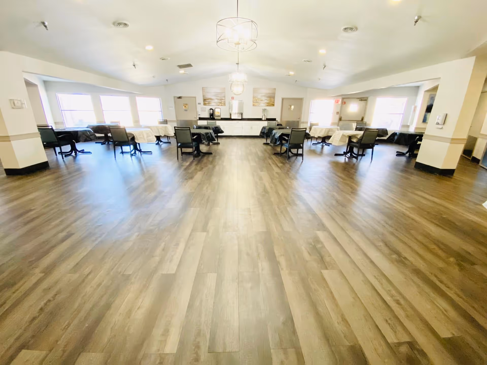 A spacious dining room with wooden flooring, multiple tables covered with white tablecloths, and black chairs arranged around them. The room is well-lit with large windows on both sides and modern ceiling light fixtures. There is a counter with coffee machines and artwork on the far wall.
