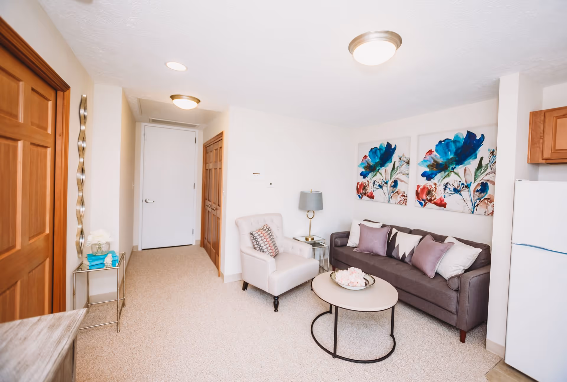 A cozy living room area with a gray sofa adorned with multiple pillows, a light beige armchair with a patterned cushion, a round coffee table with decorative items, and two large floral paintings on the wall. The room has beige carpet flooring, wooden doors, a small side table with a lamp, and a white refrigerator partially visible on the right side.