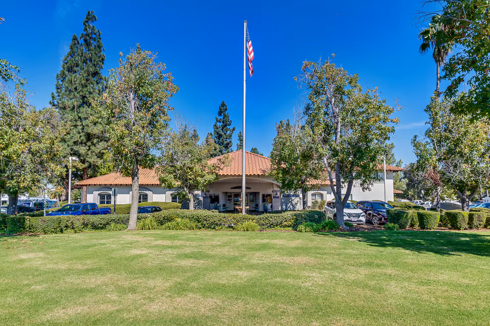 Exterior view of a single-story building with a red tile roof, surrounded by trees and bushes. A flagpole with an American flag stands in front of the building, which is set behind a well-maintained lawn. Several parked cars are visible on either side of the building under a clear blue sky.