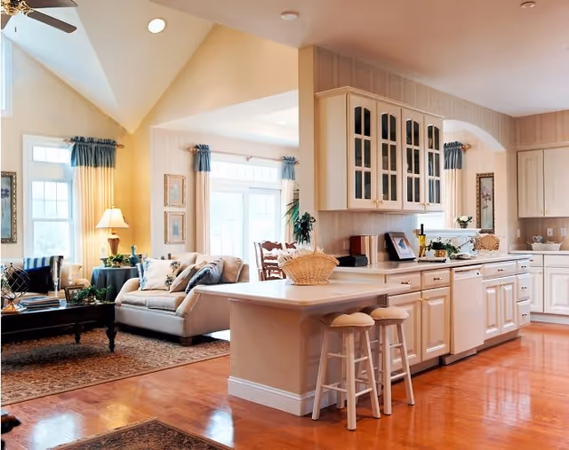 Bright and spacious open-concept living area featuring a kitchen with white cabinetry, a countertop island with two stools, and a living room with a beige sofa, armchair, side table with lamp, and large windows with blue curtains allowing natural light to fill the space.