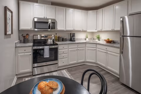 Bright L-shaped kitchen with white cabinets, stainless steel appliances, granite countertops, and a small round table with pastries.
