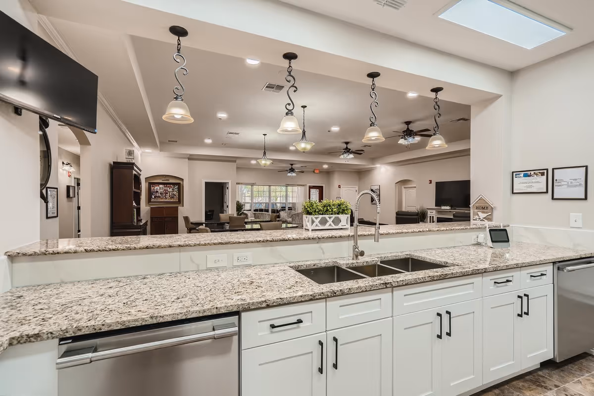 A modern kitchen area with a granite countertop, double sink, white cabinets, and stainless steel dishwasher. The kitchen opens into a spacious living room with multiple ceiling fans, pendant lights, and comfortable seating. The walls are painted light beige, and there is a large flat-screen TV mounted on the wall.
