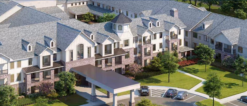 Aerial view of Harper's Station Yorktown, showing a large multi-story building with a covered entrance, surrounded by landscaped green lawns, trees, and a parking area with several cars.