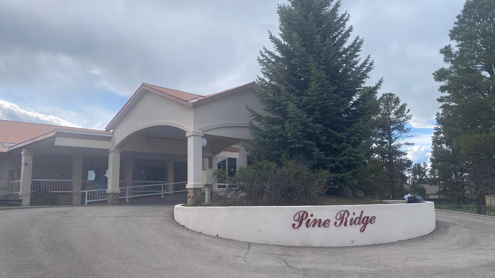 Front entrance of a low-rise building with a covered porte-cochère, circular planter and the sign "Pine Ridge" in front of tall evergreen trees.