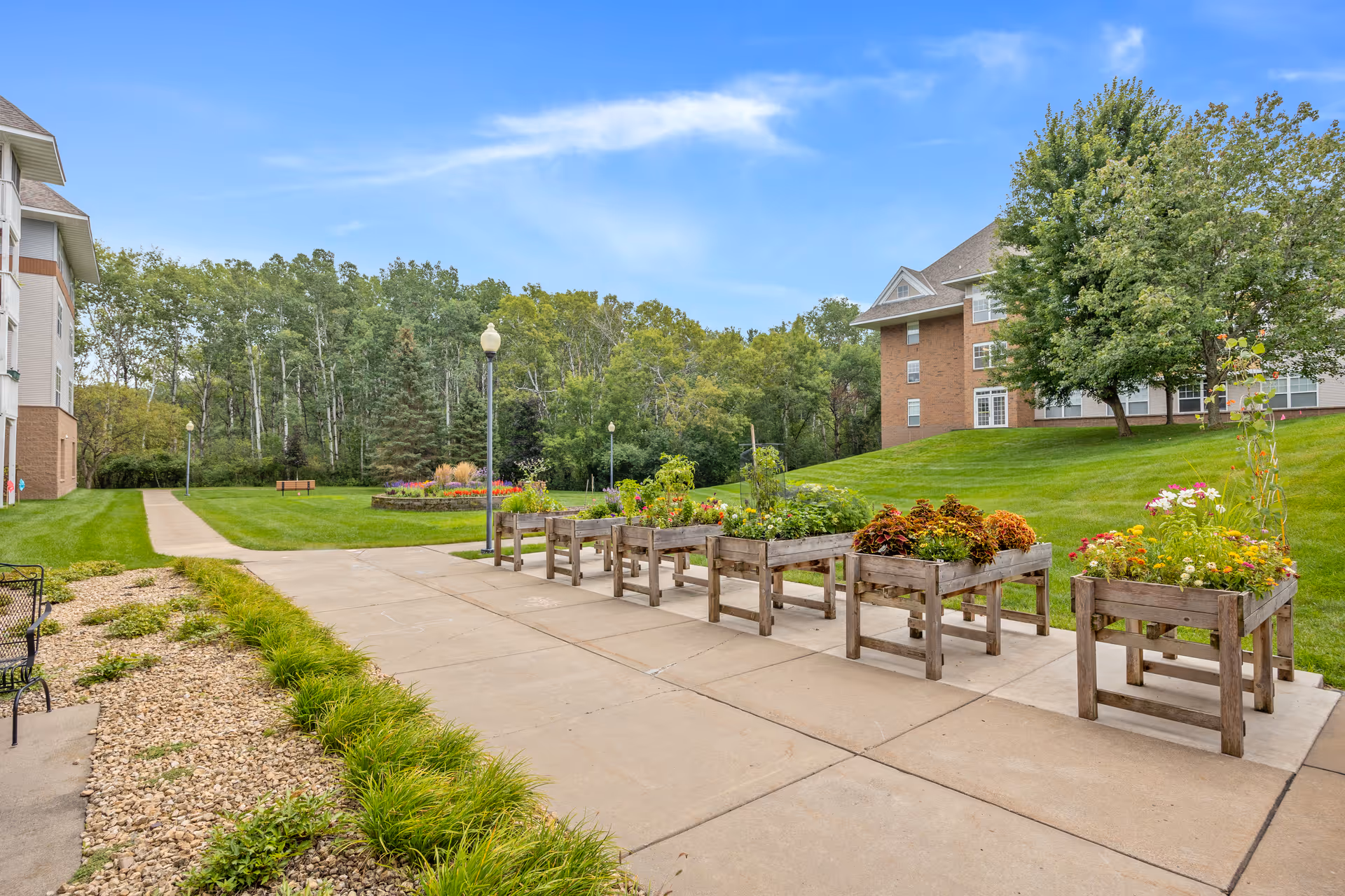 Outdoor garden area at St. Andrew’s Village featuring raised wooden planter boxes with various flowers and plants, a paved walkway, green grass, trees, and residential buildings in the background under a blue sky.