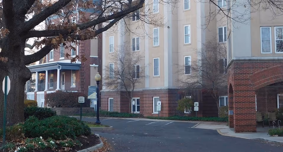Exterior view of a multi-story senior living facility building with brick and beige walls, surrounded by trees and landscaping. There is a driveway and parking area in front of the building with signs and a street lamp visible.