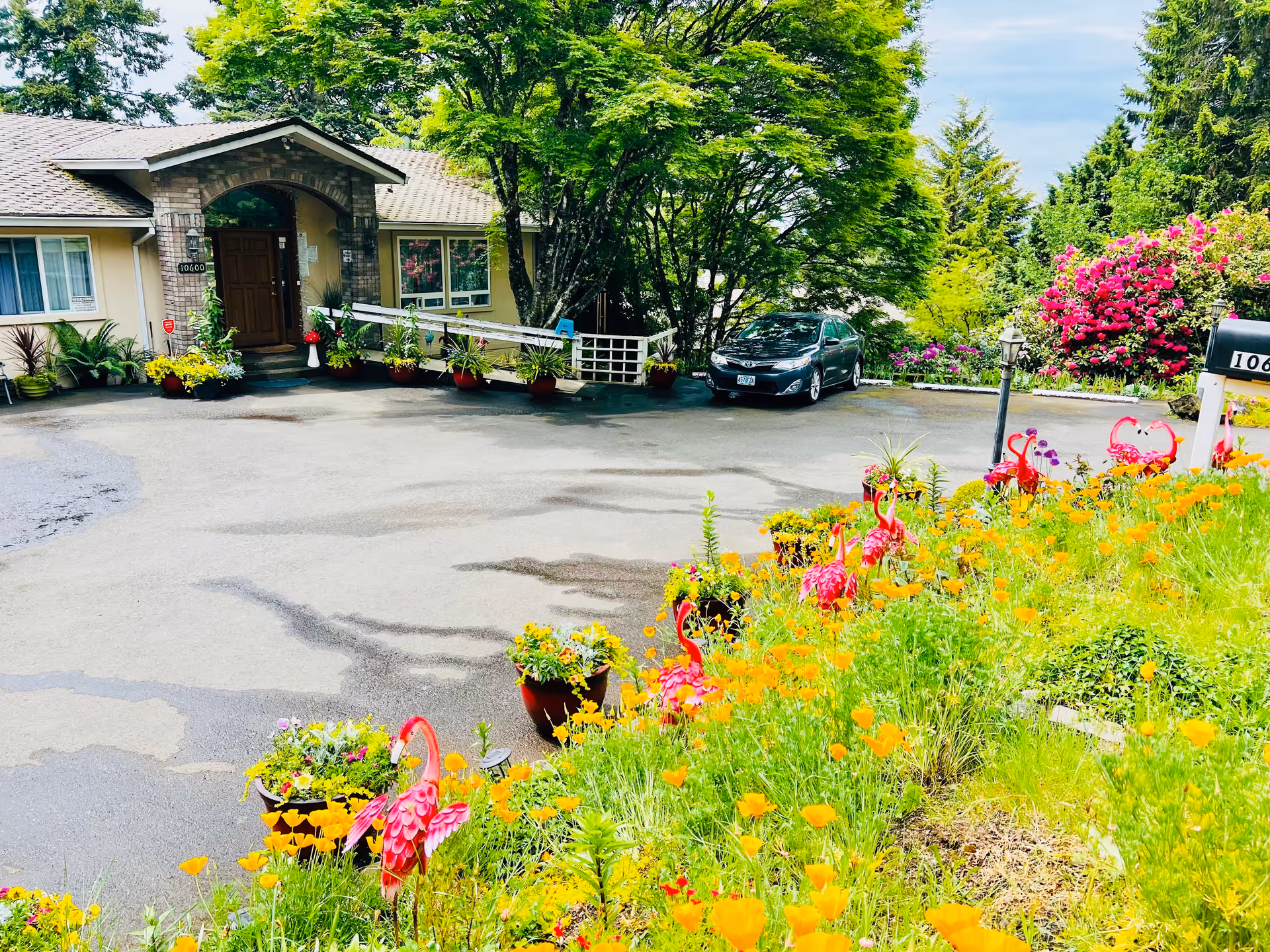 Front exterior view of a single-story building with a brick arch entrance, surrounded by lush greenery and colorful flowers. A paved driveway leads to the entrance, with a car parked near the building. Decorative pink flamingo garden ornaments and various potted plants line the driveway and garden area.