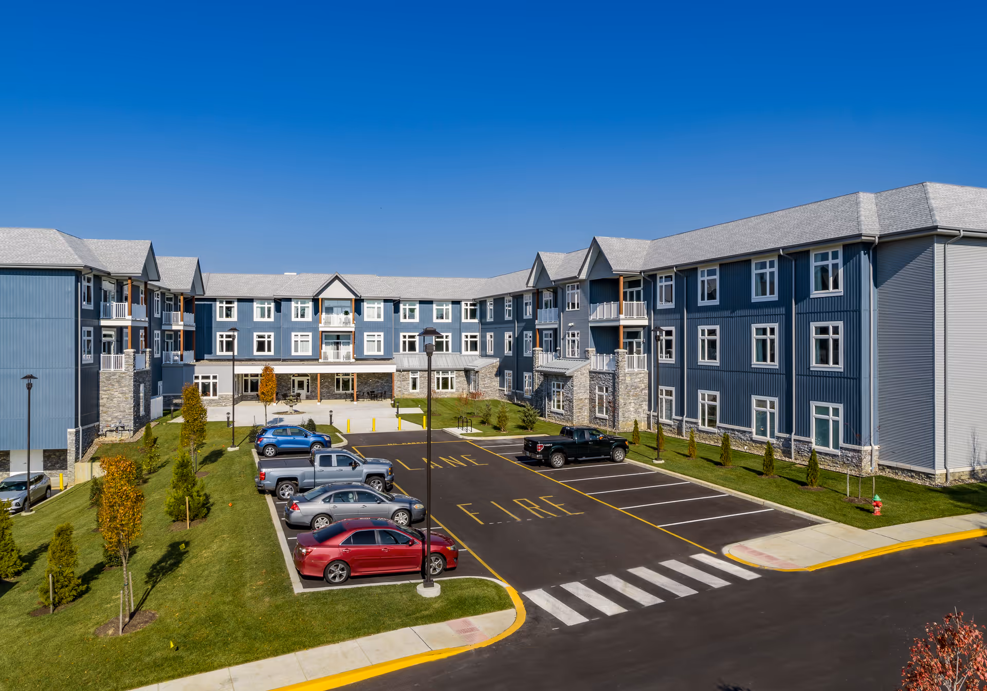 Exterior view of a three-story senior living facility building with blue siding and stone accents. The building surrounds a parking lot with several parked cars and landscaped green areas with small trees. The sky is clear and blue.