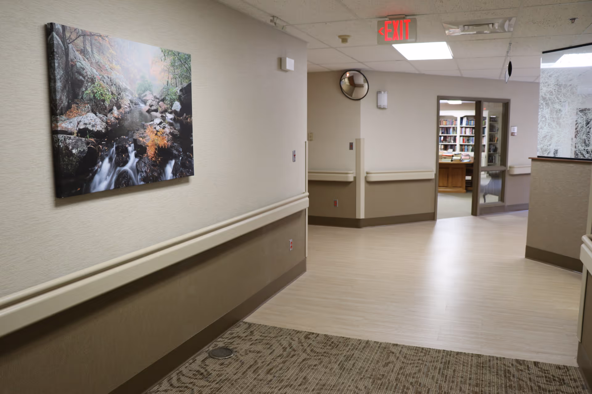 Interior hallway of a healthcare facility with beige walls and flooring. A large framed photograph of a forest stream hangs on the left wall. The hallway leads to a room with bookshelves and a wooden desk visible through glass doors. An exit sign is mounted on the ceiling.