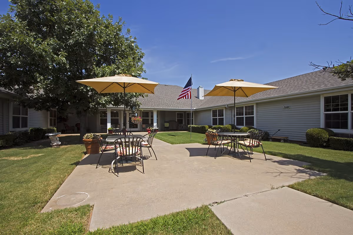 Outdoor patio area at Countryside Senior Living of Wichita Falls featuring two tables with yellow umbrellas and chairs on a concrete surface, surrounded by green grass, bushes, and a large tree. An American flag is displayed on a flagpole in the center, with single-story building walls and windows in the background under a clear blue sky.