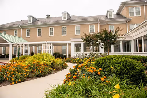 Exterior view of Glen Meadows Retirement Community showing a beige multi-story building with white trim, surrounded by a well-maintained garden with orange and yellow flowers and green shrubs along a curved walkway.