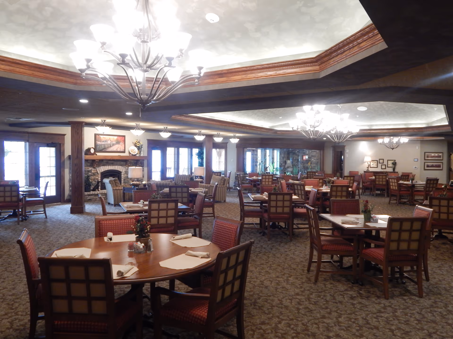 Spacious dining room with multiple wooden tables and chairs arranged neatly. The room features carpeted flooring, a decorative ceiling with recessed lighting and chandeliers, and a stone fireplace with a mantel adorned with artwork and decorative items. Large windows and glass doors allow natural light to enter the room.