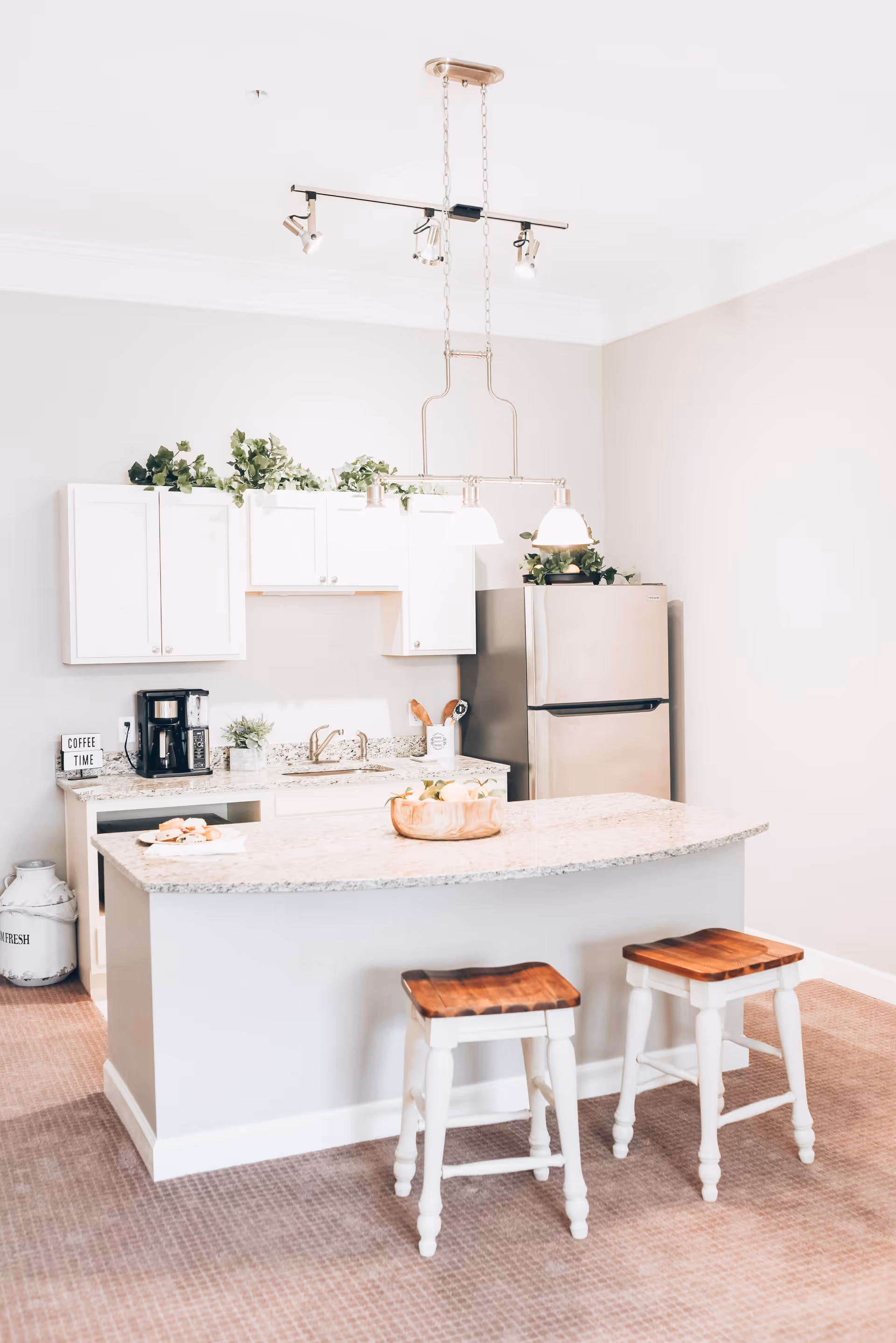 A bright kitchen area with white cabinets, a granite countertop island with two wooden stools, a stainless steel refrigerator, a coffee maker on the counter, and green plants decorating the top of the cabinets. Three pendant lights hang above the island.