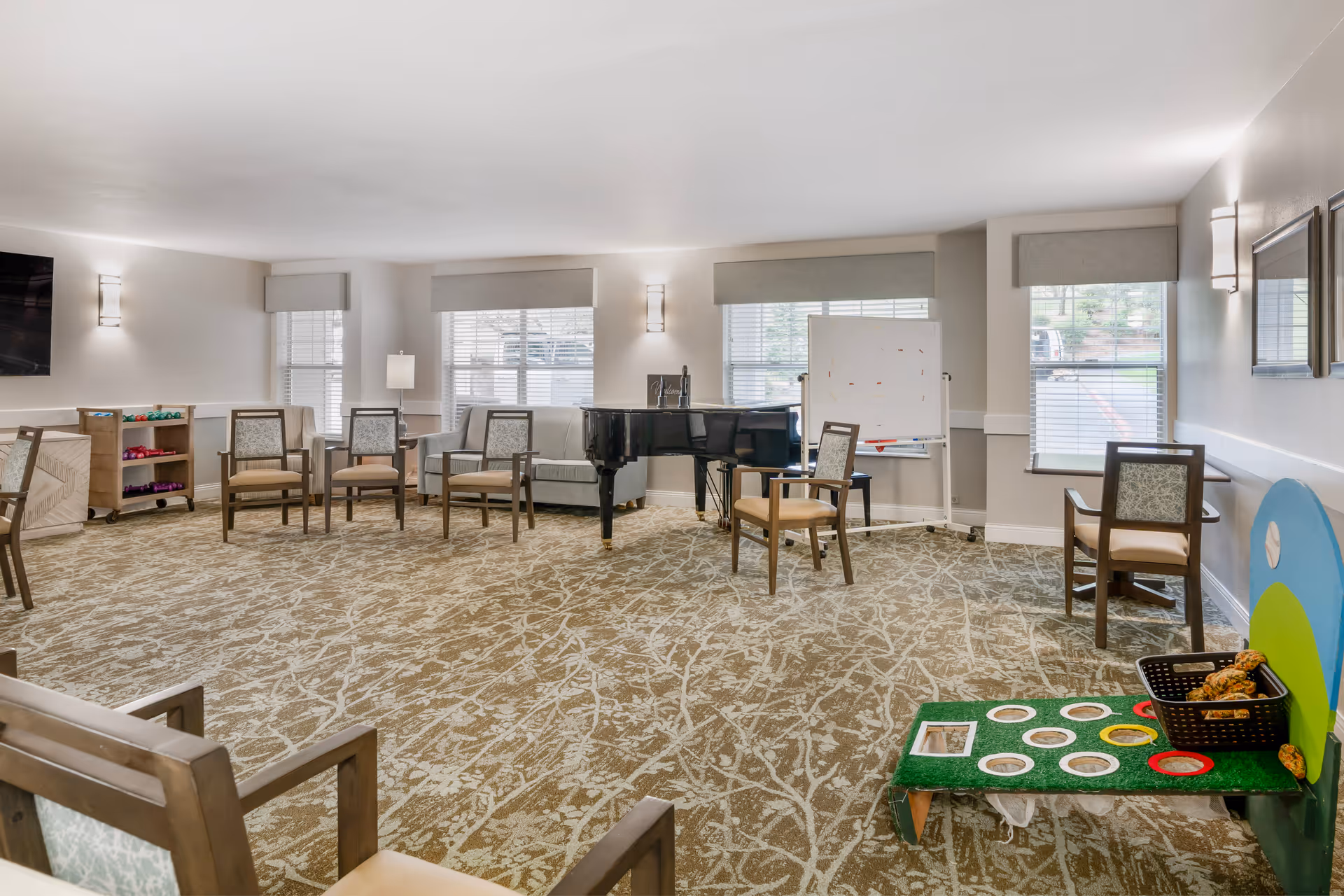 A spacious, well-lit common room with several chairs arranged in a circle, a black grand piano, a whiteboard on a stand, and a small game setup with bean bags and holes on a green board. The room has large windows with blinds, beige walls, and patterned carpet flooring.