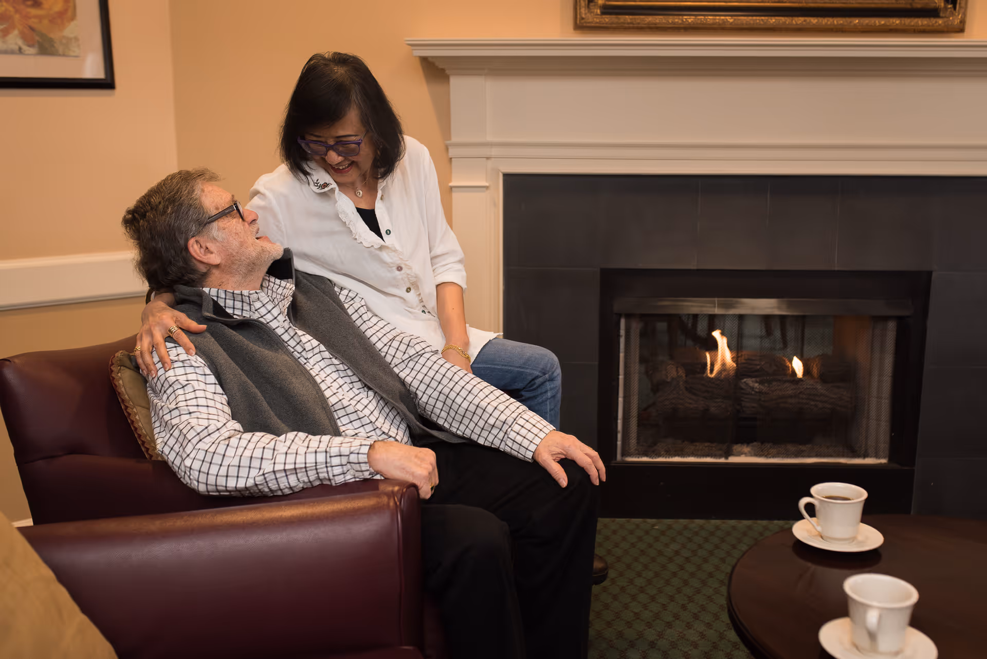 Two people sitting by a lit fireplace in a cozy lounge, smiling with coffee cups on a table.