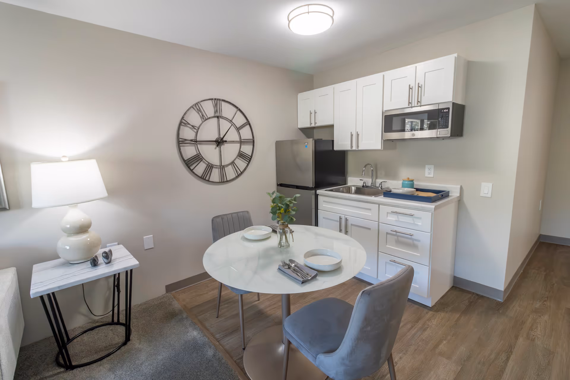 A small kitchen and dining area in a senior living facility apartment. The kitchen has white cabinets, a stainless steel refrigerator, a microwave, and a sink. In front of the kitchen is a round white dining table set with two place settings, including bowls, plates, and utensils. Two gray upholstered chairs are positioned at the table. A side table with a white lamp and a large decorative wall clock are visible on the left side. The flooring is a combination of wood and carpet.