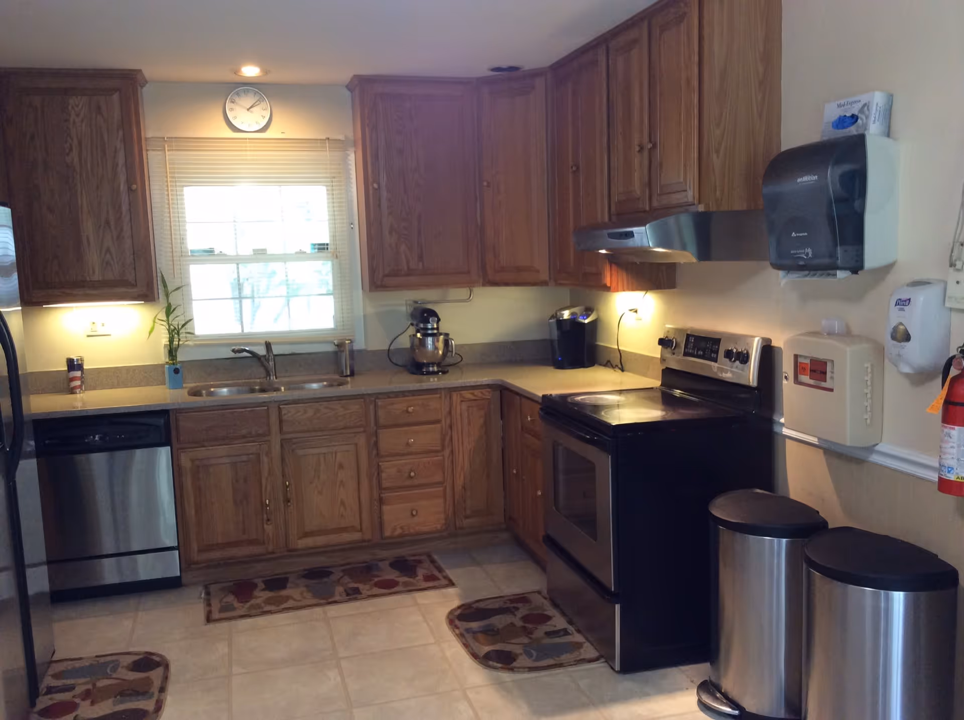 A kitchen with wooden cabinets, a stainless steel dishwasher, stove, and refrigerator. There is a window above the sink with blinds, a clock on the wall, and various kitchen appliances on the countertops. Two stainless steel trash bins and a fire extinguisher are visible on the right side.