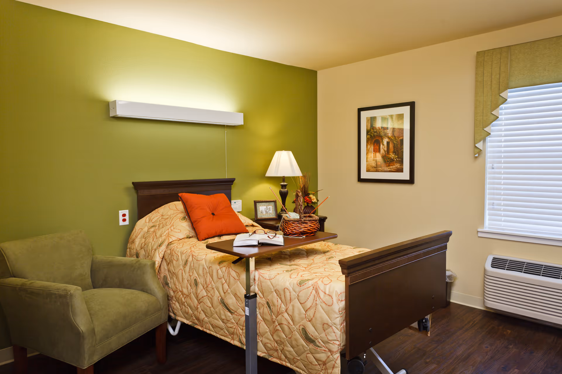 A neatly furnished senior bedroom with a single adjustable bed, overbed table, bedside lamp and decor, green armchair, and a window with blinds.