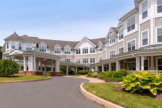 Exterior view of a large senior living facility building with multiple windows, a covered entrance driveway, and landscaped greenery including bushes and grass under a clear blue sky.