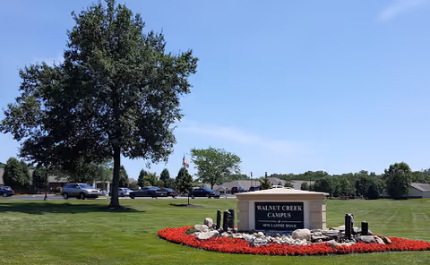 Entrance sign for Walnut Creek Campus on a manicured lawn with red flowers, trees, parked cars, and a blue sky.