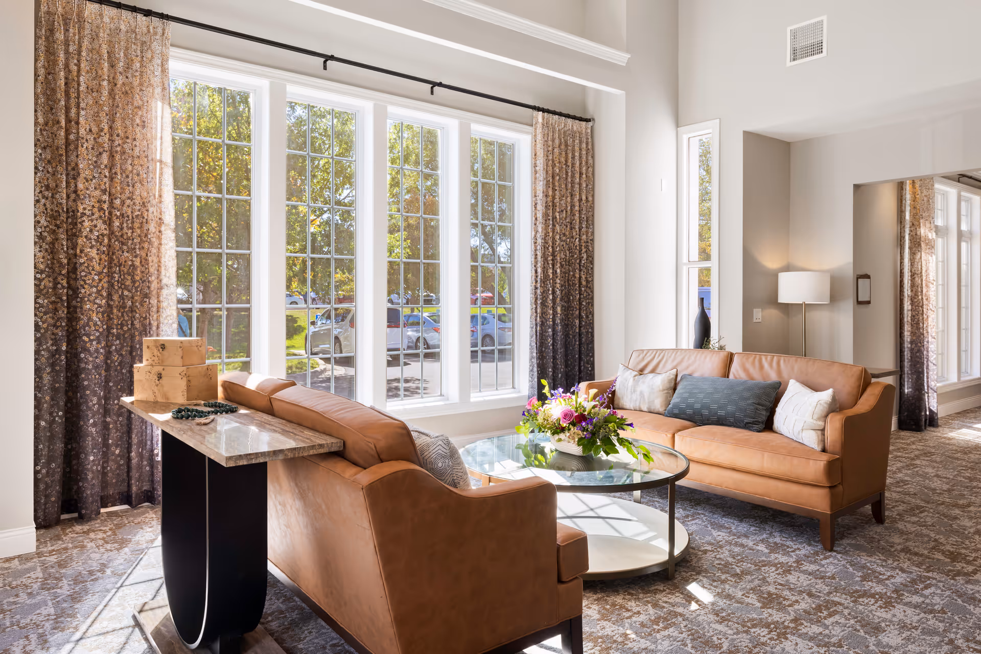 A bright and cozy living room area with two tan leather sofas facing each other around a round glass coffee table with a floral arrangement. Large windows with patterned curtains let in natural light, and a floor lamp stands in the corner. The room has a patterned carpet and light-colored walls.