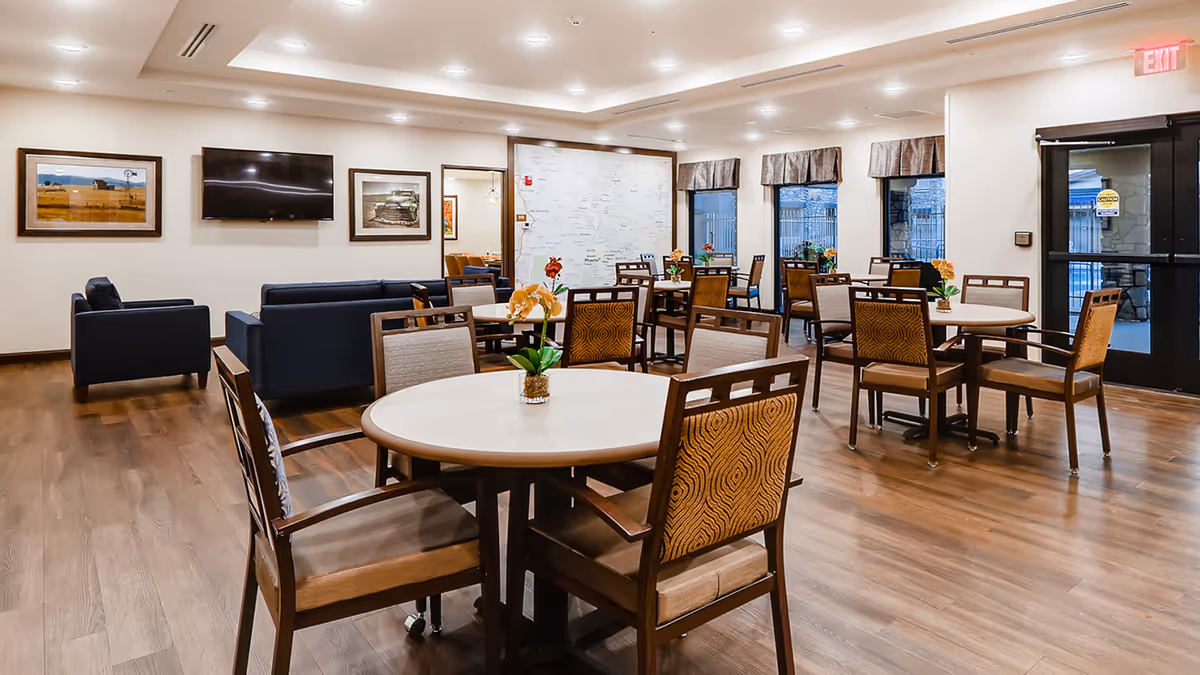 A spacious, well-lit common area with multiple round tables and chairs arranged for seating. Each table has a small flower arrangement. In the background, there are two dark blue sofas facing a wall-mounted flat screen TV, flanked by framed pictures. Large windows with valances let in natural light, and a glass door with an exit sign is visible on the right. The room has wood flooring and recessed ceiling lights.