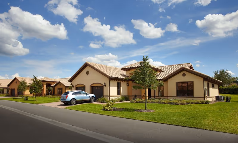 Exterior view of a single-story residential building with a tiled roof, beige walls, and a driveway with a parked silver SUV. The building is surrounded by a well-maintained lawn and small trees under a partly cloudy blue sky.