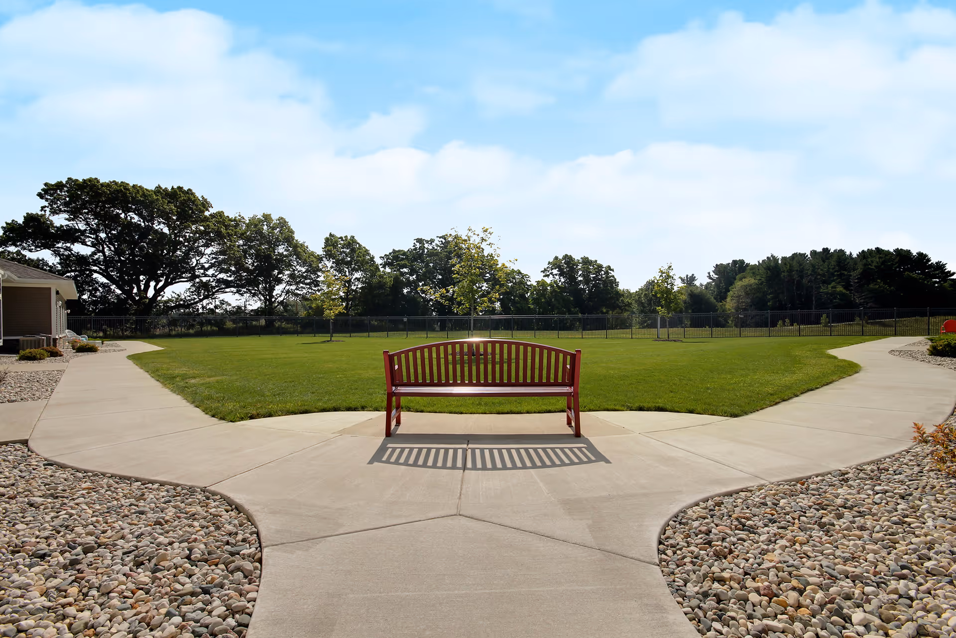 A red bench sits on a concrete pathway that splits into two directions, surrounded by green grass and landscaped areas with small rocks. Trees and a fence are visible in the background under a partly cloudy sky.