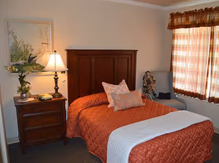 A cozy bedroom with a single bed covered in an orange quilt and two decorative pillows. Next to the bed is a wooden nightstand with a lamp, a small plant, and a phone. A framed artwork hangs on the wall above the nightstand. There is a window with plaid curtains allowing natural light into the room, and a cushioned chair is placed near the window.