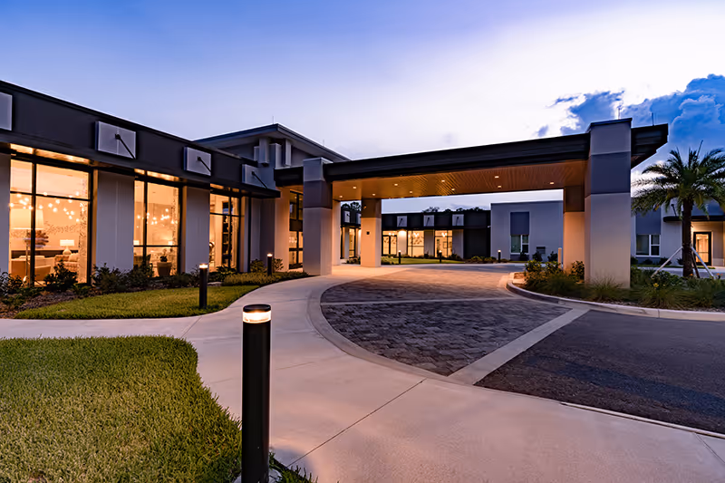 Dusk view of the Village on the Green front entrance showing a covered porte-cochère, curved driveway, landscaped lawn, and illuminated interior windows.