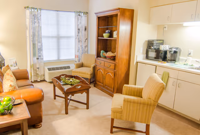 A cozy living room area with a brown leather sofa, two striped armchairs, a wooden coffee table with a decorative bowl, a wooden cabinet with shelves, and a kitchenette with a microwave, coffee maker, and sink. A window with floral curtains lets in natural light.