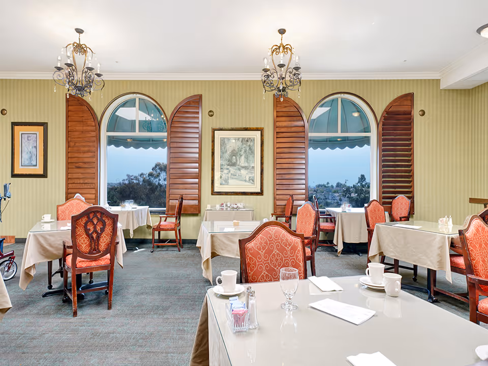 Formal dining room with tables and upholstered chairs, arched windows with wooden shutters and chandeliers.