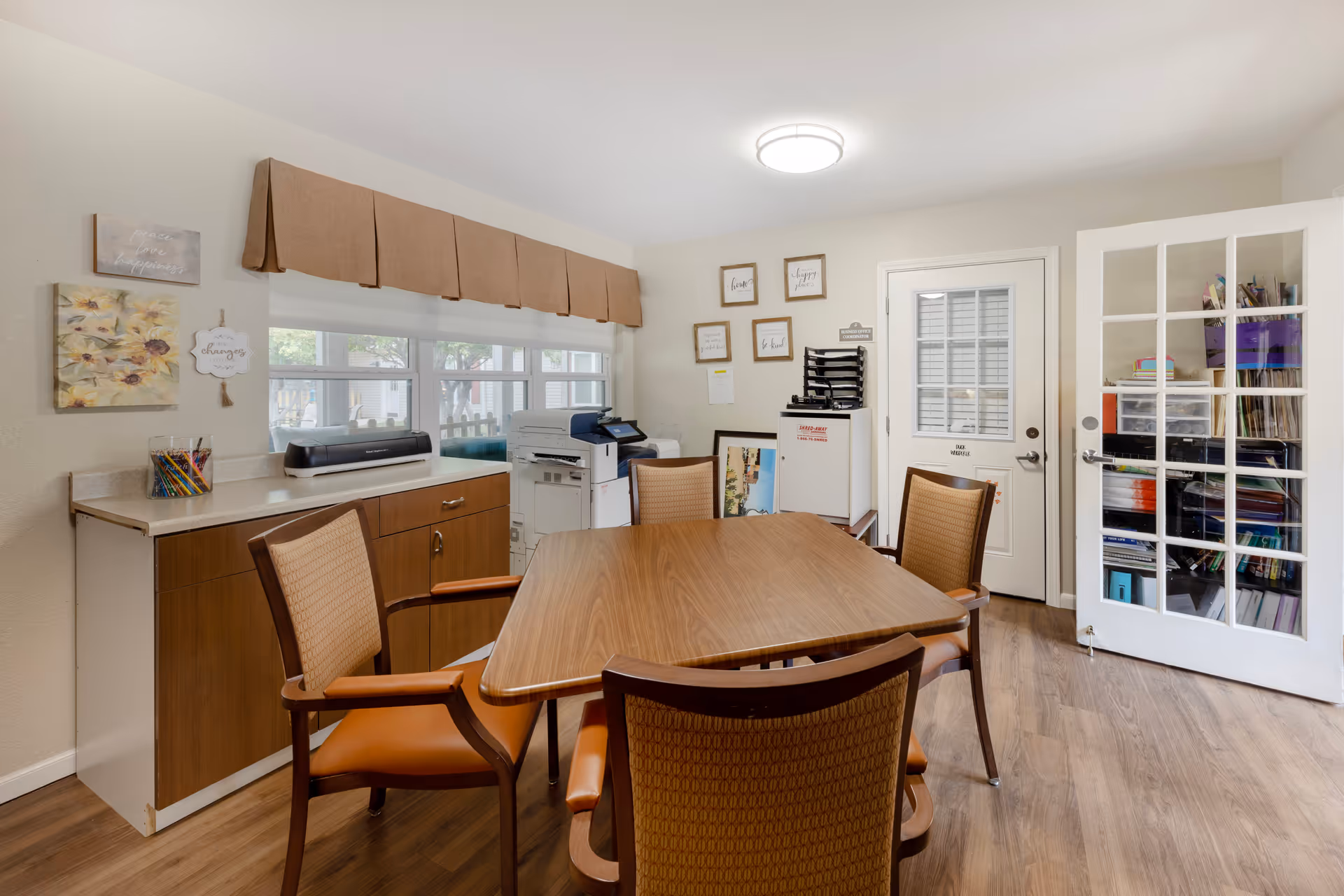 A small meeting or office room with a wooden table surrounded by four chairs with orange cushions. There is a countertop with a printer and colored pencils on the left side under a window with a brown valance. On the right side, there is a white door with glass panels and a shelving unit filled with office supplies. The walls have framed inspirational quotes and artwork. The floor is wooden, and the room is well-lit with a ceiling light.