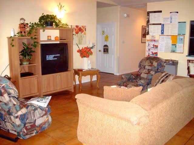 A cozy living room area with a beige couch and two patterned armchairs facing a wooden entertainment center with a TV. The TV screen displays a message. There are plants and decorative items on the entertainment center. A small wooden side table with a vase of flowers is next to a white door with a yellow decoration. Various notices and papers are pinned on a bulletin board on the wall.