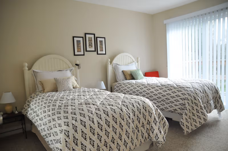 A bright bedroom with two twin beds featuring white headboards and patterned bedspreads. Each bed has multiple pillows. There are two small bedside tables with lamps, and three framed pictures hang on the wall above the beds. A large window with vertical blinds lets in natural light.