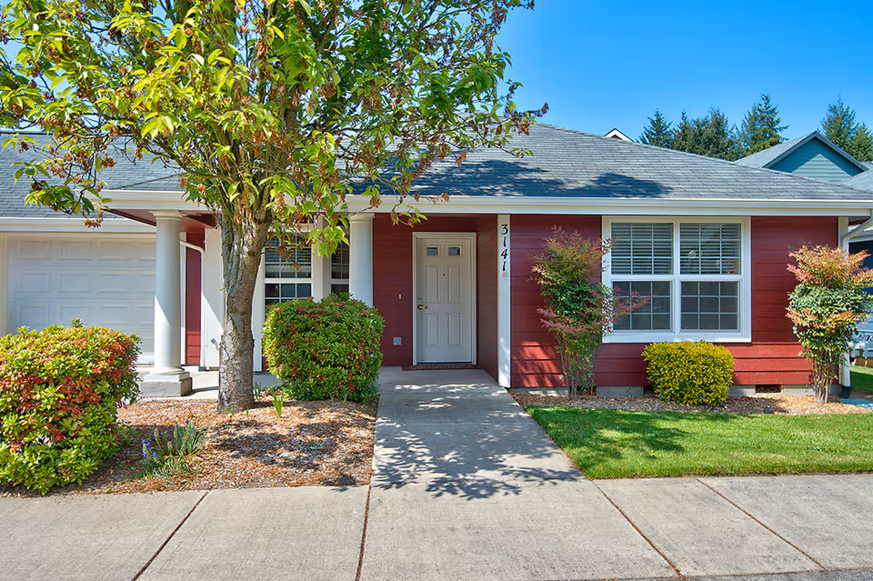 Front exterior of a single-story red residence with a covered porch, white-trimmed windows, garage, and landscaped yard.