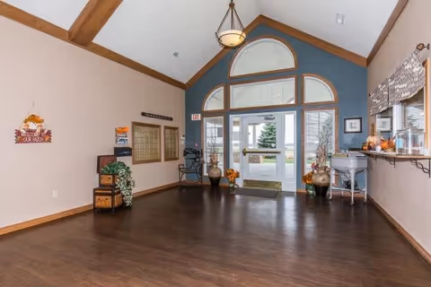 Interior view of a spacious entryway or lobby area with wooden floors and high ceilings featuring exposed wooden beams. The far wall has a large glass door with decorative windows above, allowing natural light to enter. There are decorative plants and vases on either side of the door, a small table with autumn-themed decorations on the right, and a wall-mounted mailbox area on the left. A sign on the left wall reads 'Welcome to Our Place'.