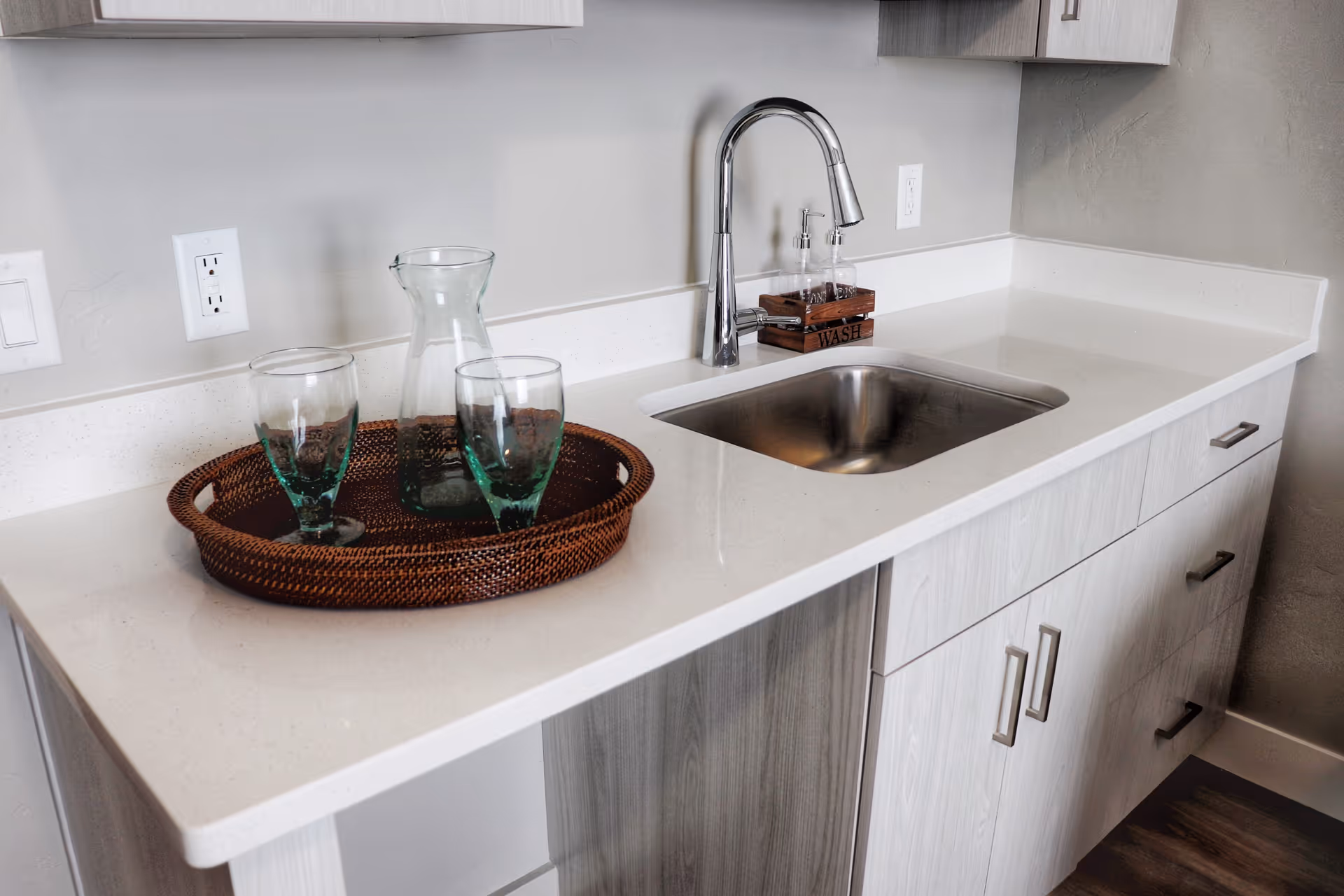 A modern kitchen countertop with a stainless steel sink and a chrome faucet. On the countertop, there is a brown woven tray holding a glass pitcher and two drinking glasses. There is also a wooden soap dispenser holder labeled 'WASH'. The cabinetry below is light wood with silver handles.