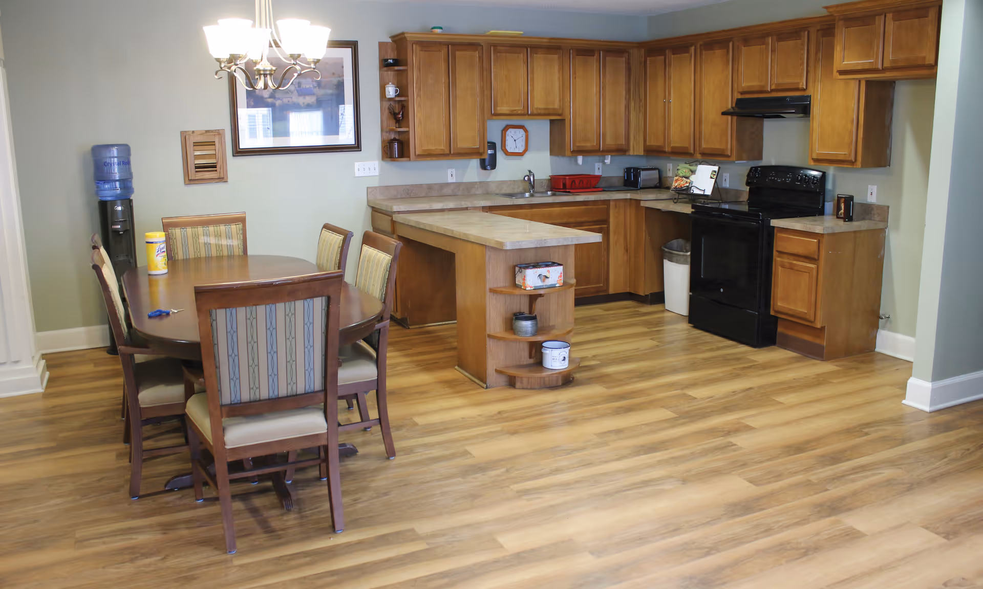 A kitchen and dining area in a senior living facility with wooden cabinets, a black stove, a countertop with an island, and a dining table with six chairs. There is a water cooler and a chandelier above the dining table. The floor is wood-patterned.