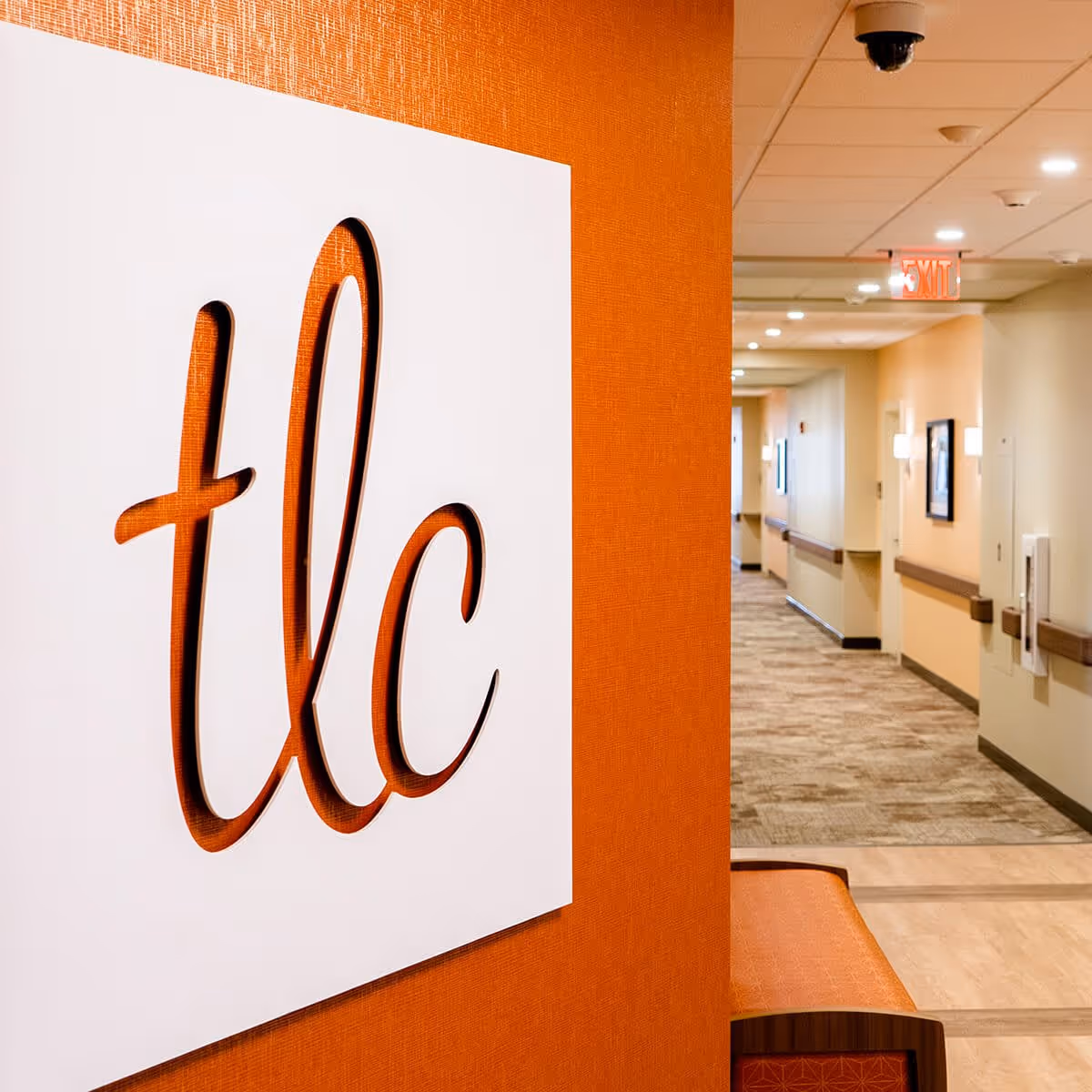 Interior hallway of TLC Living Community with an orange wall featuring a white sign with the letters 'tlc' cut out. The hallway has beige walls, carpeted floor, ceiling lights, and an exit sign at the end.