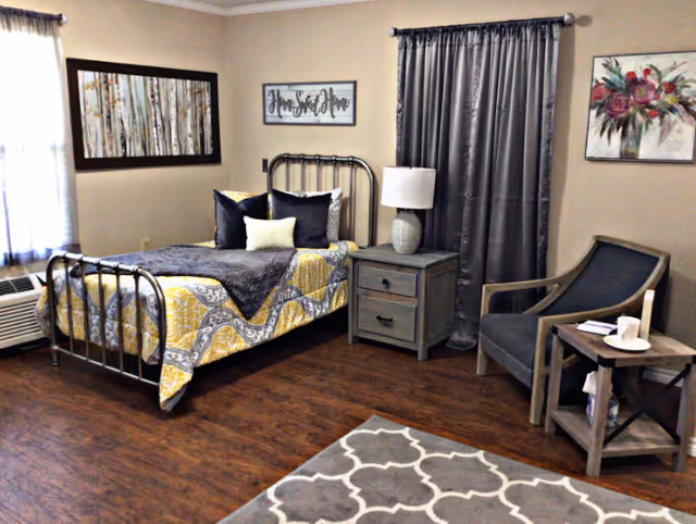 A cozy bedroom with a metal-framed bed covered in a yellow, gray, and white patterned quilt and multiple pillows. Next to the bed is a wooden nightstand with a white lamp. There is a gray curtain covering a window, a framed floral painting on the wall, and a framed picture with the text 'Home Sweet Home'. A gray armchair and a small wooden side table with a cup and book are also visible. The floor is wooden with a gray and white patterned rug.