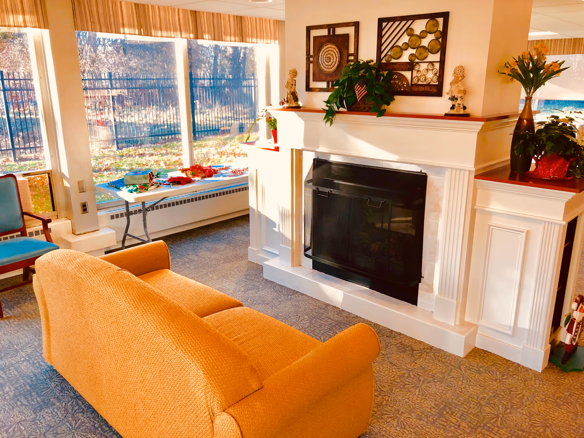A cozy senior living community lounge area with a mustard yellow sofa facing a white fireplace. The fireplace mantel is decorated with plants, small statues, and framed artwork. Large windows behind a table with holiday decorations let in natural light and show a fenced outdoor area.