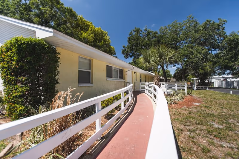 Exterior view of a single-story yellow building with a white roof and windows, featuring a curved red walkway with white railings leading to the entrance. The area is surrounded by green trees, bushes, and grass under a clear blue sky.