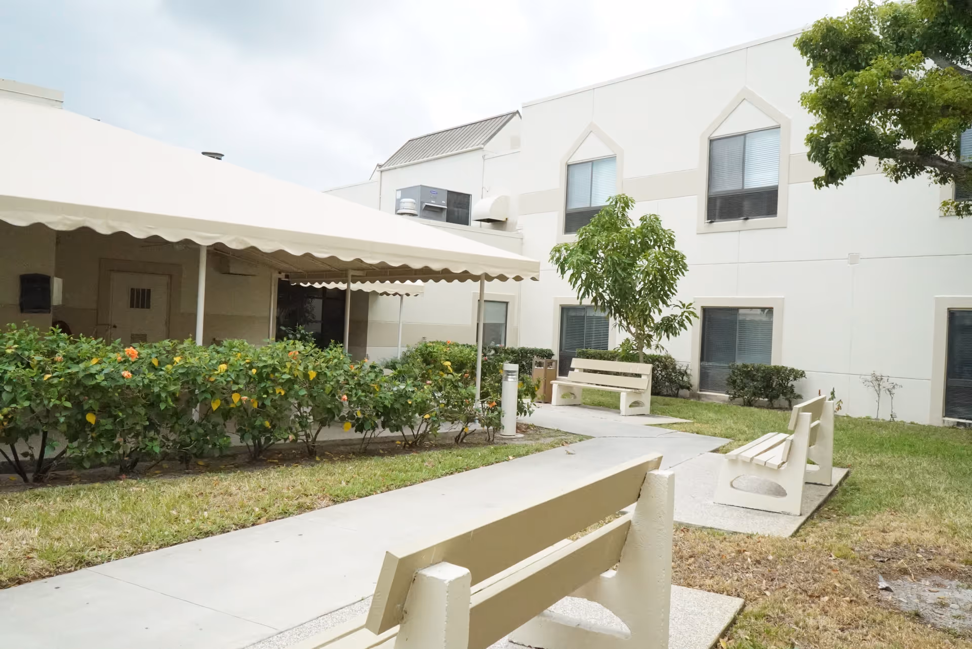 Outdoor courtyard area at Palm Garden of West Palm Beach featuring beige benches along a paved walkway, green bushes with orange flowers, a small tree, and a white building with multiple windows in the background under a cloudy sky.