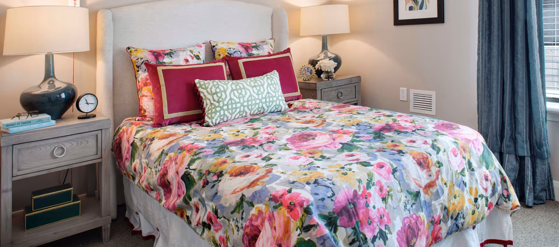A cozy bedroom with a bed featuring a floral-patterned bedspread and multiple decorative pillows in shades of pink, red, and green. There are two bedside tables, each with a lamp, and one table has books, a clock, and glasses. A framed picture hangs on the wall, and a window with blue curtains is visible on the right side.