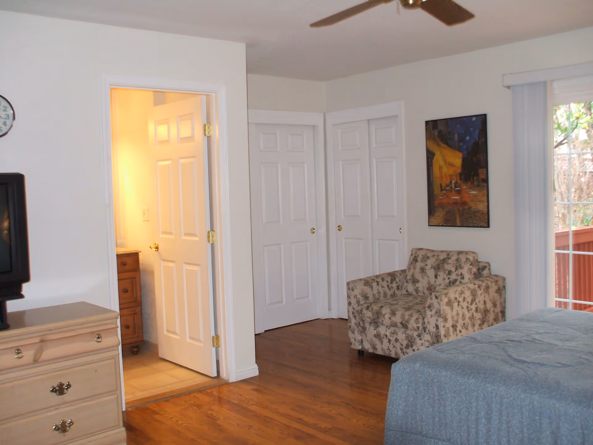 A bedroom with hardwood floors featuring a floral armchair, a bed with a blue bedspread, a wooden dresser with a TV on top, and an open door leading to a bathroom. There are two closed white closet doors, a ceiling fan, a wall clock, a framed painting, and a sliding glass door with vertical blinds that opens to an outdoor area.