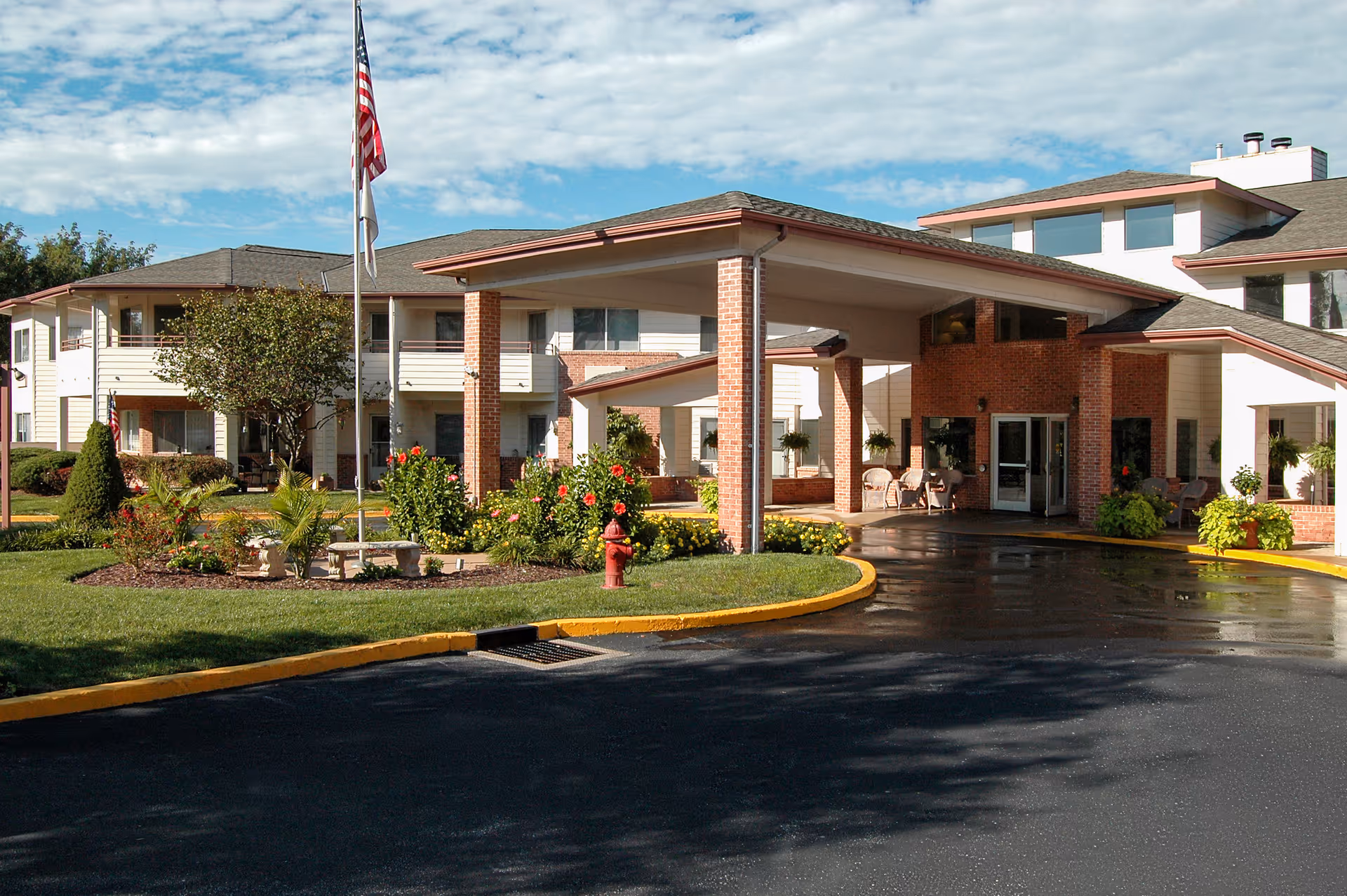 Exterior view of Holiday Briarcrest Estates senior living facility showing a covered entrance with brick pillars, landscaped garden with flowers and shrubs, an American flag on a flagpole, and a multi-story building with balconies and large windows under a partly cloudy sky.