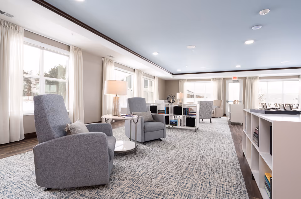 A bright and spacious senior living common area with large windows letting in natural light. The room features comfortable gray armchairs with small side tables and lamps, a patterned carpet, and white shelving units filled with books and decorative items. The walls are painted in neutral tones, and the ceiling is light blue with recessed lighting. There is a door at the far end of the room leading outside.